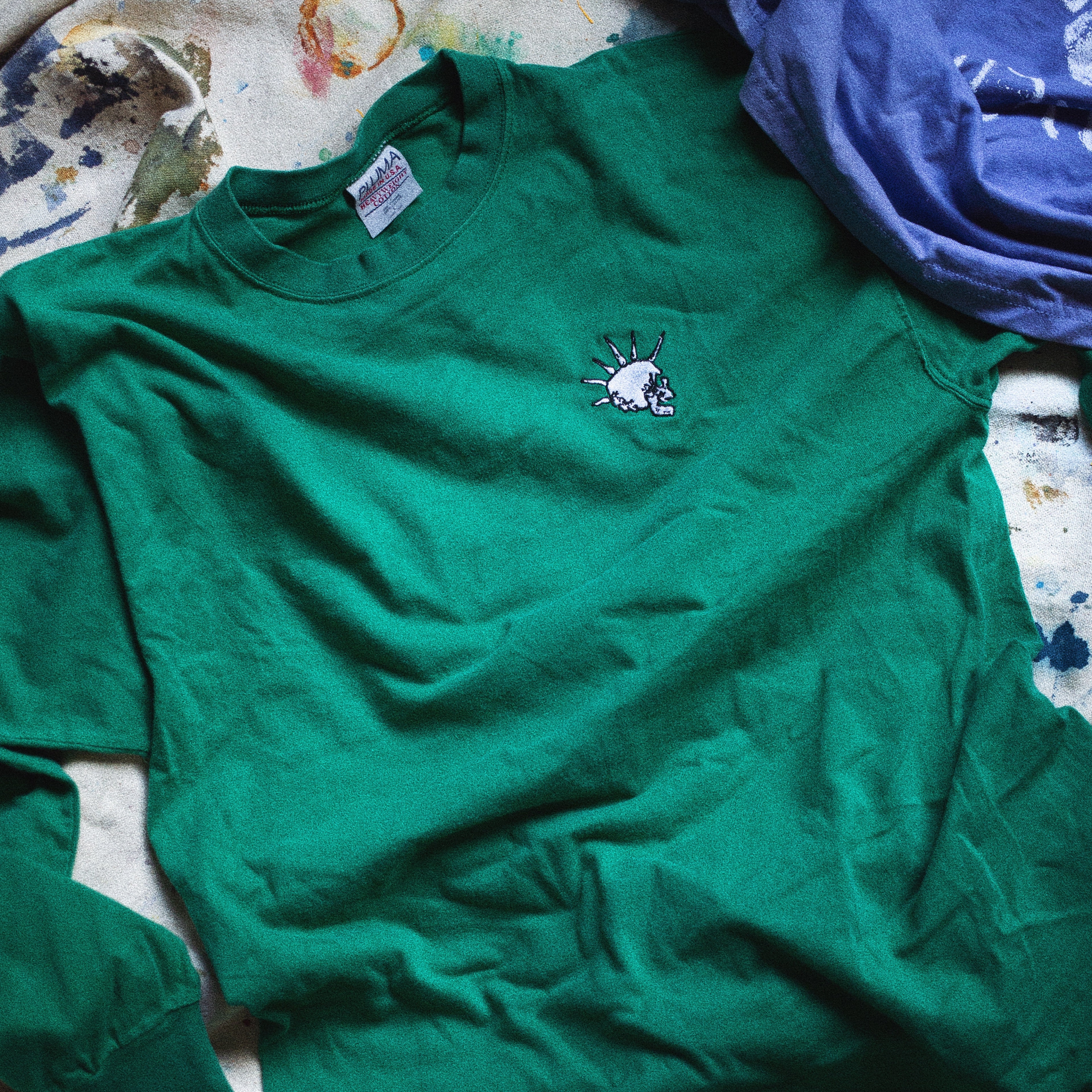 Green longsleeve shirt with skull logo on a canvas with colorful stains and paint
