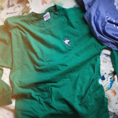 A green crewneck long-sleeved t-shirt with a small skull graphic on the left chest, laid flat on a paint-splattered surface next to a lavender shirt.
